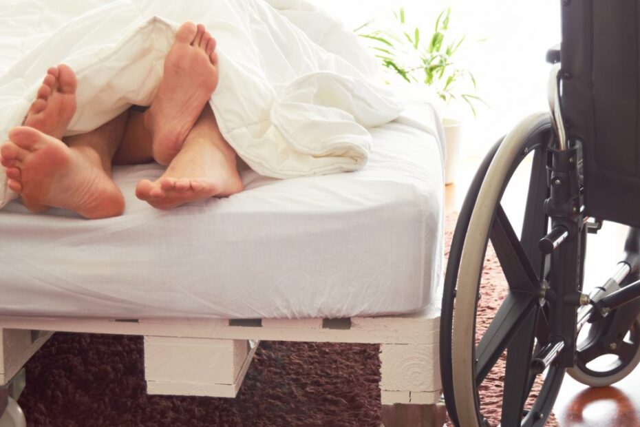 Afbeelding van een bed waarin onder de witte deken vier voeten te zien zijn. Naast het bed staat een rolstoel.