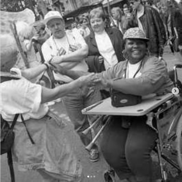 Zwartwit foto van een lachende Zwarte vrouw in een rolstoel die in de camera kijkt. Ze houdt de handen vast van een witte vrouw met kort haar en een rugzak. Ze dansen samen in de straten van Amsterdam (fotograaf: Marian Bakker)
