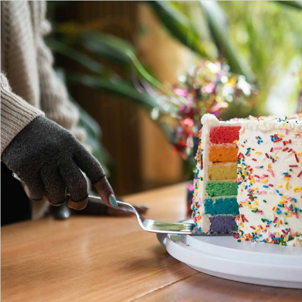 een bruine hand met lichtblauwe nagels pakt een stuk taart dat rood, oranje, geel, groen, blauw en paars is aan de binnenkant en wit met gekleurde spikkels aan de buitenkant.