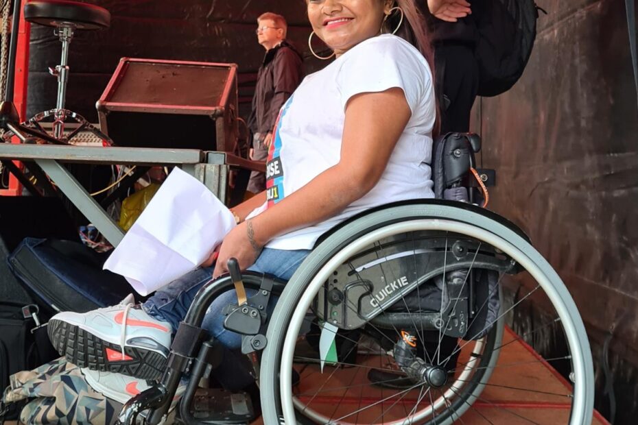 Foto van Chedda, een bruine vrouw met lang bruin haar. Ze is te zien vanaf de zijkant. Ze heeft een wit t-shirt aan en zit in haar rolstoel. Op de achtergrond is een podium te zien.